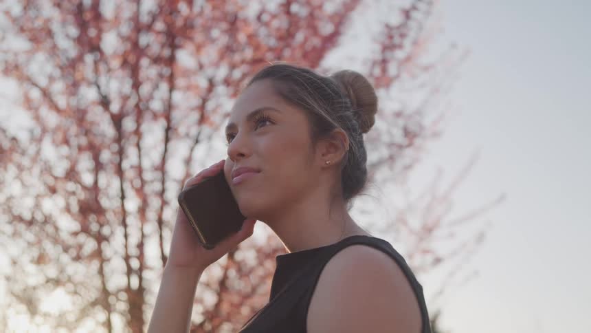 A woman standing outside talks on her cell phone next to a large tree with fall leaves