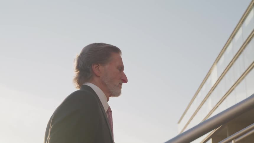 Senior man wearing a suit and necktie walks up the stairs of an office building at sunset