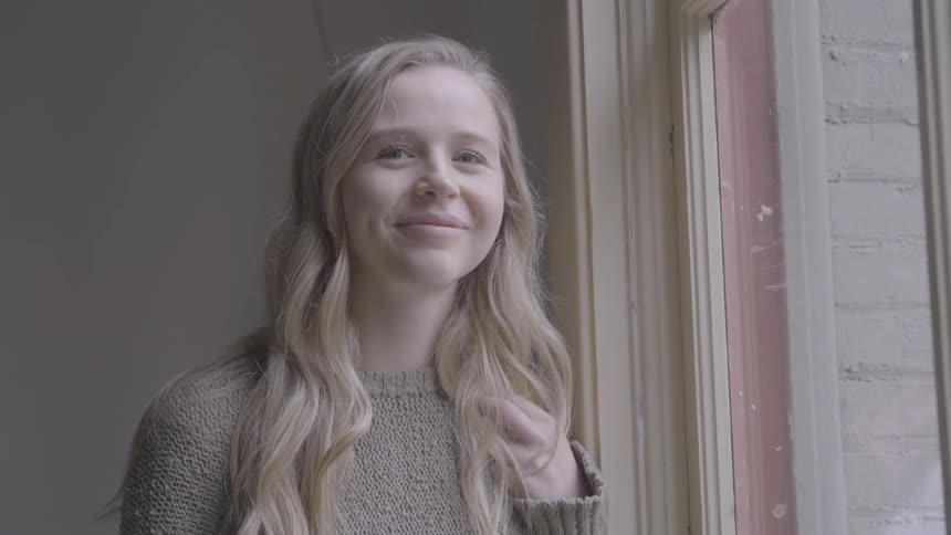 Young blonde woman next to a window turns to look at the camera smiling