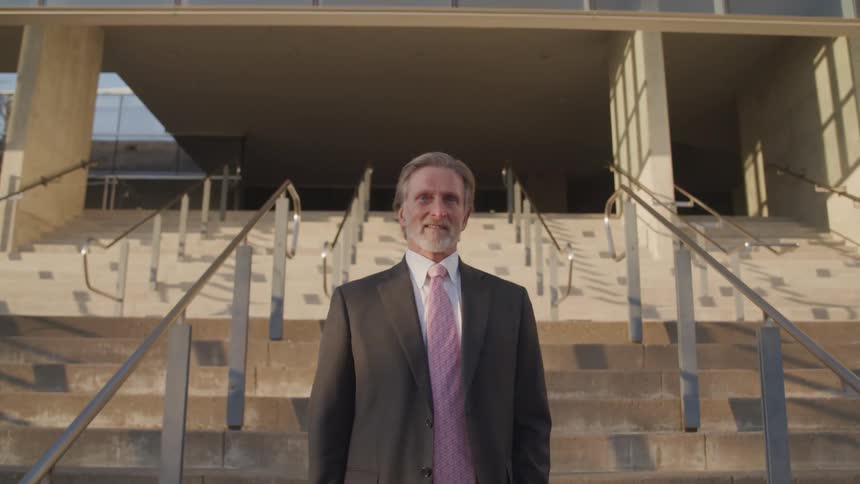 Businessman with a beard and a suit stands and smiles in front of the office building stairs