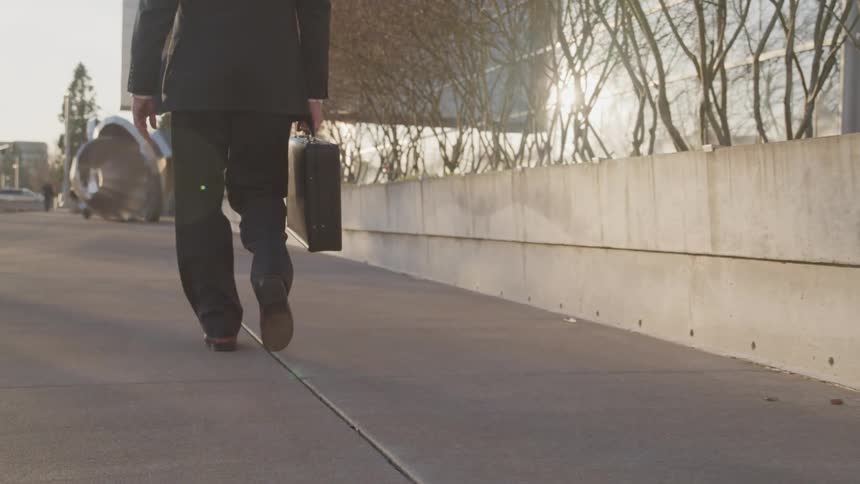 Man wearing a suit and holding a briefcase walks next to a building in city downtown at sunset