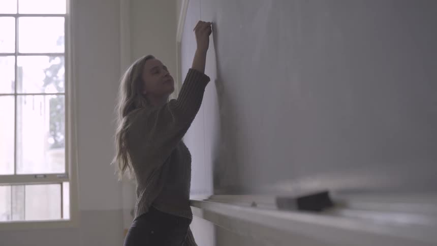 Blonde girl with a grey pullover writes on a blackboard with chalk in the class