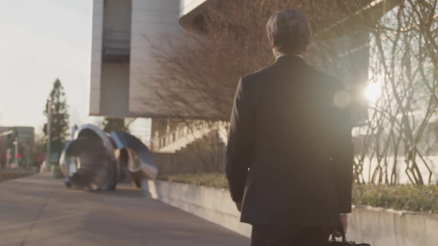 A man in a business suit walks along a sidewalk towards a building with a sculpture in front of it