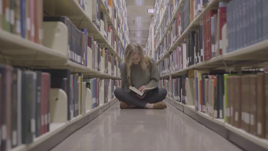 A student sits on the floor in library stacks and reads a book