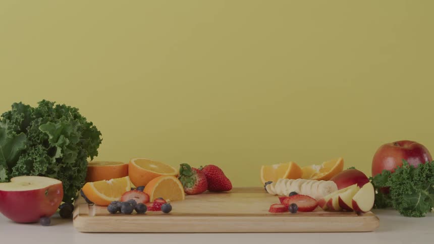 A person taking a blender full of fruit and vegetables from a cutting board on a table surrounded by fruit and vegetables
