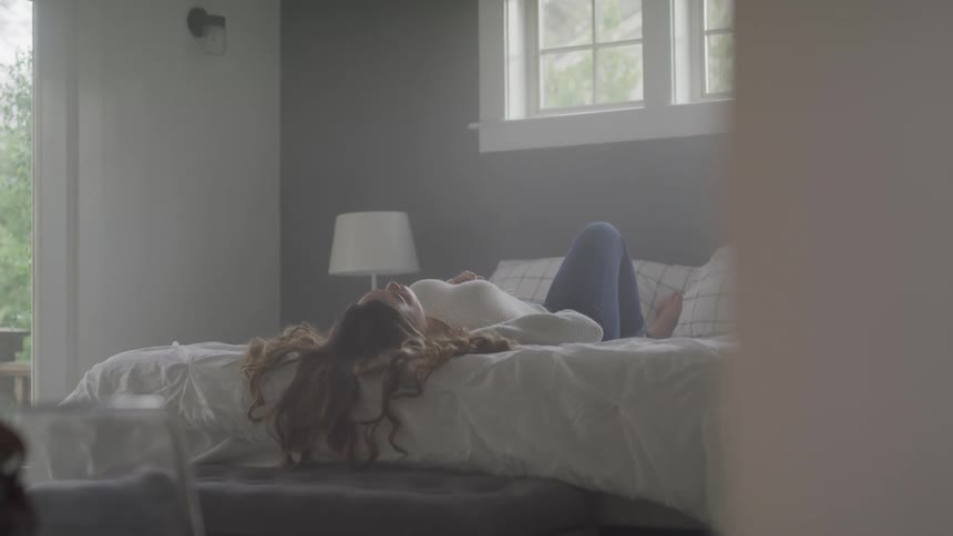 Woman with a white sweater and jeans lies down on bed in bedroom
