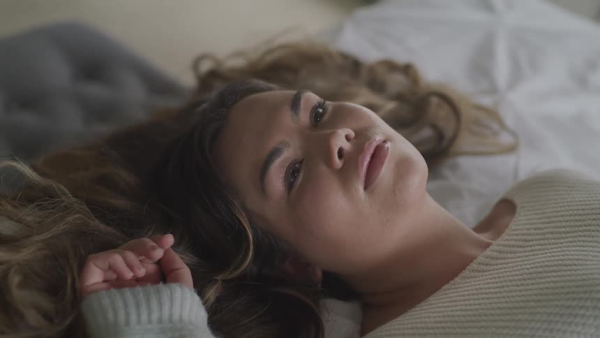 Woman with brown wavy hair lies down on bed and closes her eyes