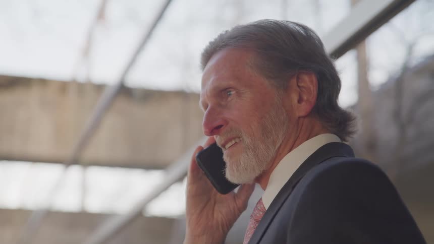 Side view of a businessman with a grey beard a suit talking on a smart phone outside of an office building
