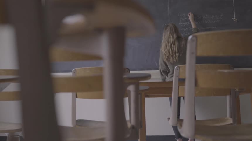 Blonde girl with a grey pullover writes on a blackboard with chalk in the classroom
