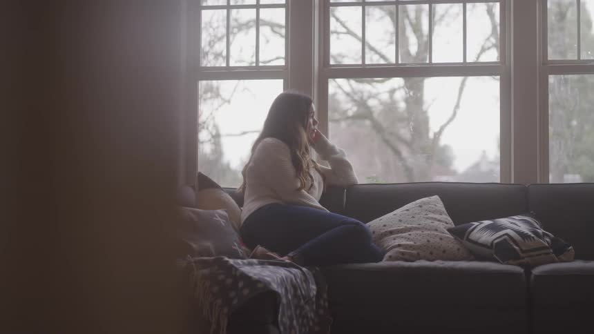 A girl sits sideways on a couch and looks out the window of her home looking a bit sad