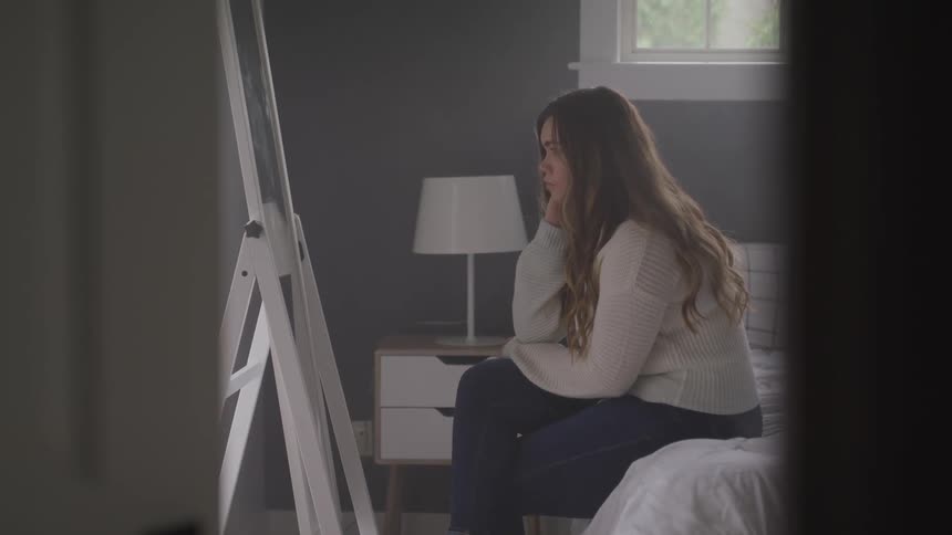 A teenager sits on her bed and looks at herself in a full length mirror