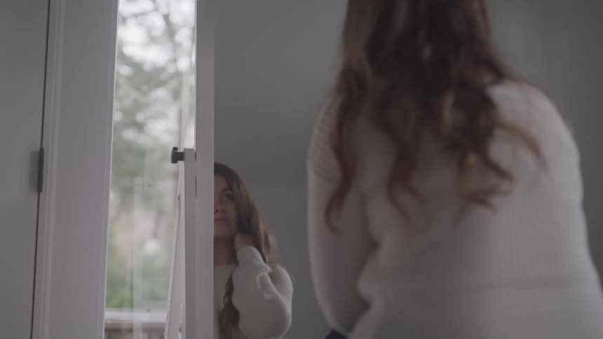 Woman with brown wavy hair sitting on a bed looks in a mirror