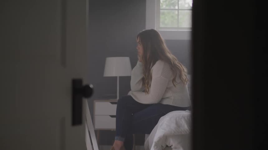 Woman with brown hair a white sweater and jeans sits on bed and looks in the mirror in bedroom