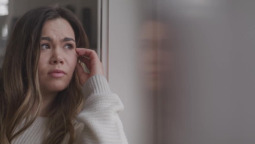 A woman sitting near a window gazes out of it contemplatively