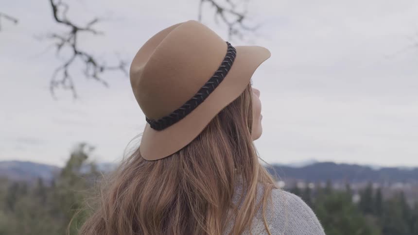 A woman wearing a wool camel hat stands outside overlooking a forest with a mountain range in the distance