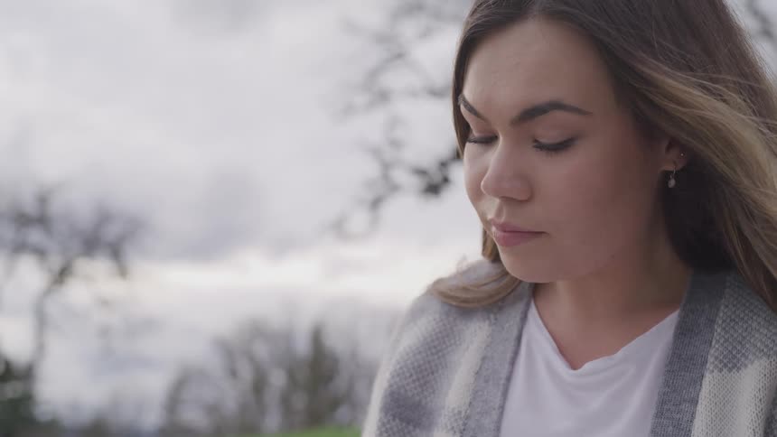 Woman with brown hair and a grey sweater sits on green grass and reads a book on a cloudy day