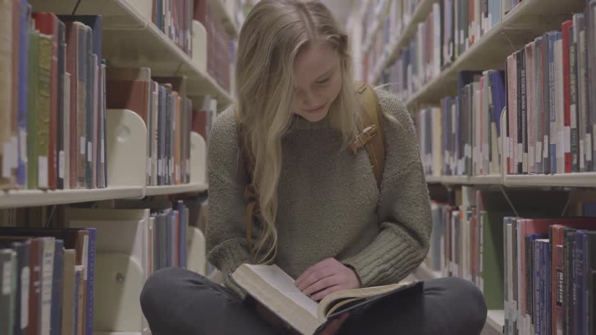 A young woman wearing a backpack sits in the aisle of a library while reading a book