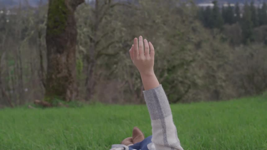 Woman laying on her back over the grass raises her hand to the sky