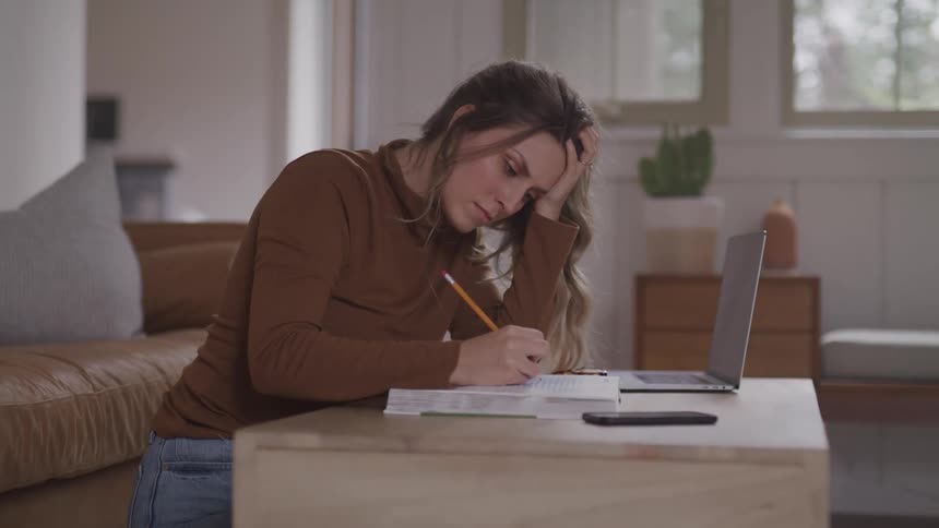 A woman kneels at a coffee table writing in a book with a laptop beside her