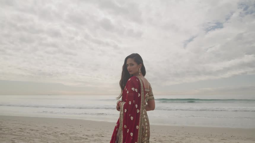 Woman with red saree stands on the white sand beach next to the ocean on a cloudy day