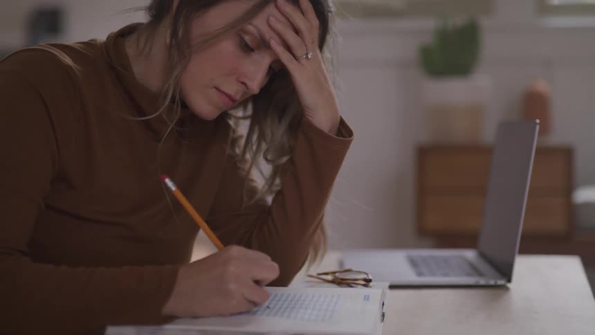 Woman with a brown sweater rests her head on her hand and writes on a paper