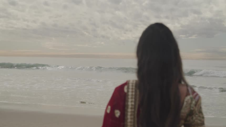 Woman with red traditional dress stands on white sand beach next to the ocean on a cloudy day