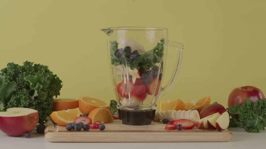 A person sprinkles berries and fruit into a blender sitting on a cutting board