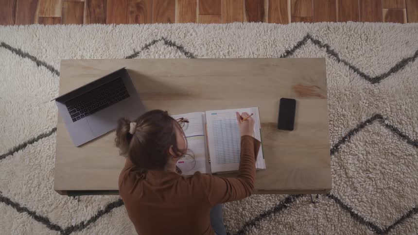 A woman sitting by a desk with a laptop and a smart phone on it while writing in a book