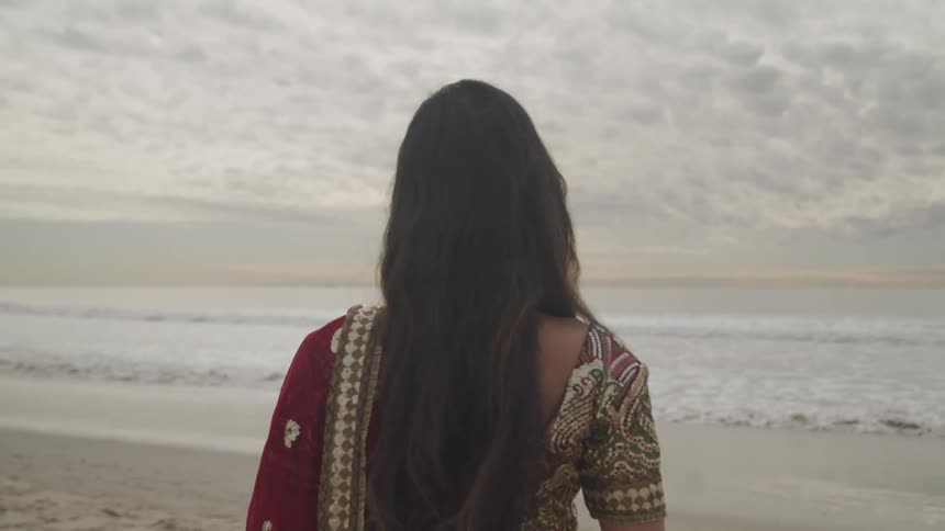 An indian woman in a saree gazes out at the sea while standing on a beach on a cloudy day