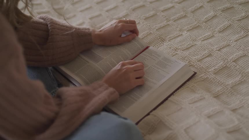 Woman with a brown sweater and jeans sits on a bed and reads a book in bedroom