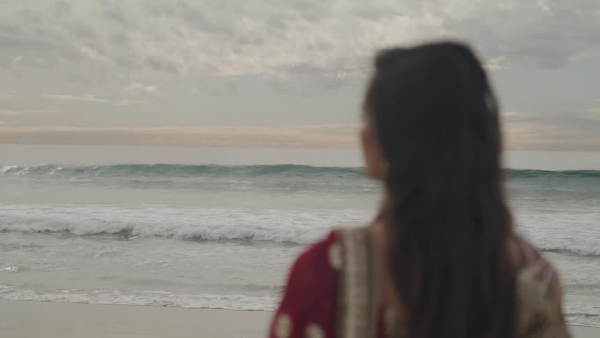 Woman with red traditional dress stands on white sand beach next to the ocean on a cloudy day