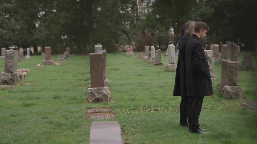 A man and a woman solemnly stand side by side in a graveyard during the day
