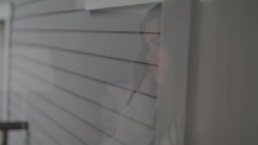 A womans reflection is shown in a window as she sits on her bed with her head in her hand