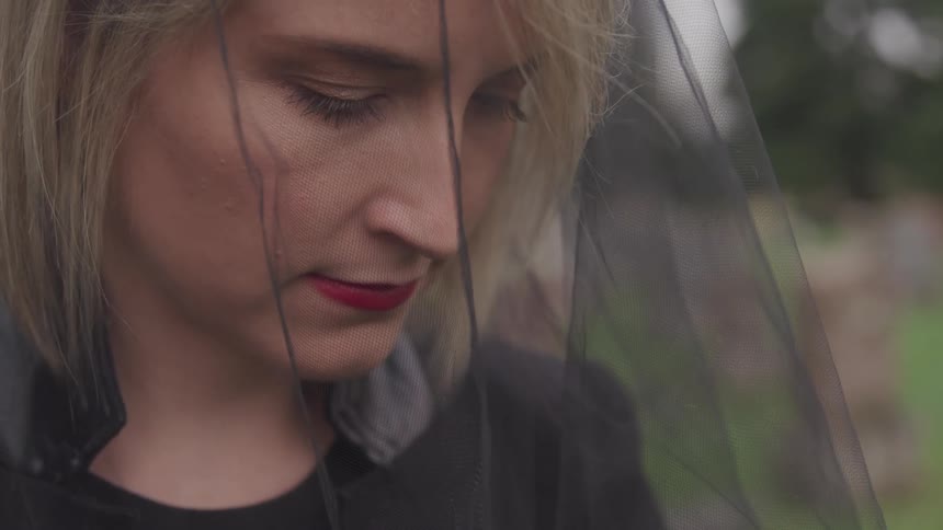 Blonde woman with makeup and in a black veil looks down in graveyard