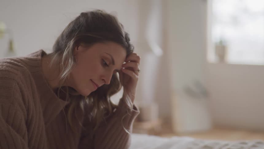 Woman with brown hair and a brown sweater sits on a bed and looks down in bedroom