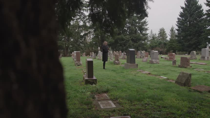 A woman standing in a cemetery surrounded by trees with the sky in the background