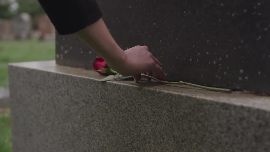 A woman places a rose on top of a grave in a cemetery at midday