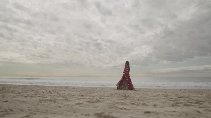 Woman with red traditional dress walks on white sand beach next to the ocean on a cloudy day