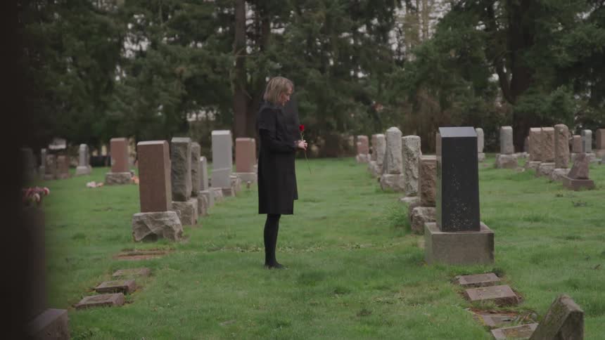 A woman dressed in all black holds a rose while standing solemnly in front of a large headstone in a cemetery