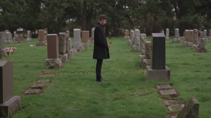 Young man holding a rose mourns in front of a grave in the cemetery