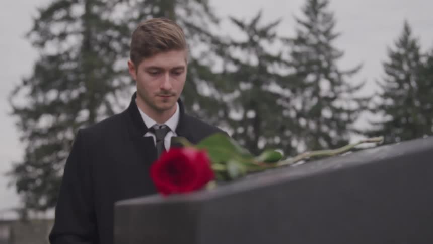 A man grieves as he stands in front of a headstone with a rose on top