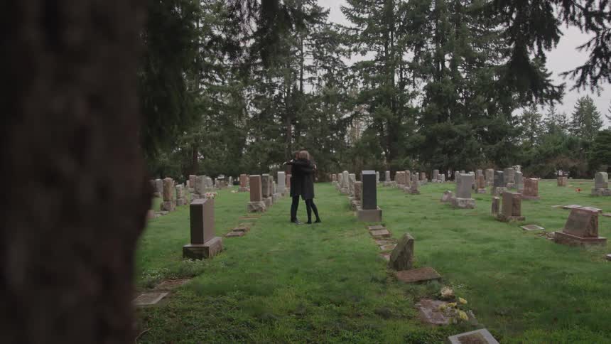 Mourners embrace standing between graves in cemetery bordered by conifer trees