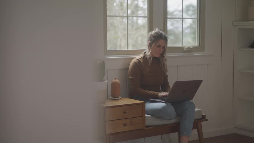 Woman with a brown sweater and brown hair sitting in an armchair uses a grey laptop
