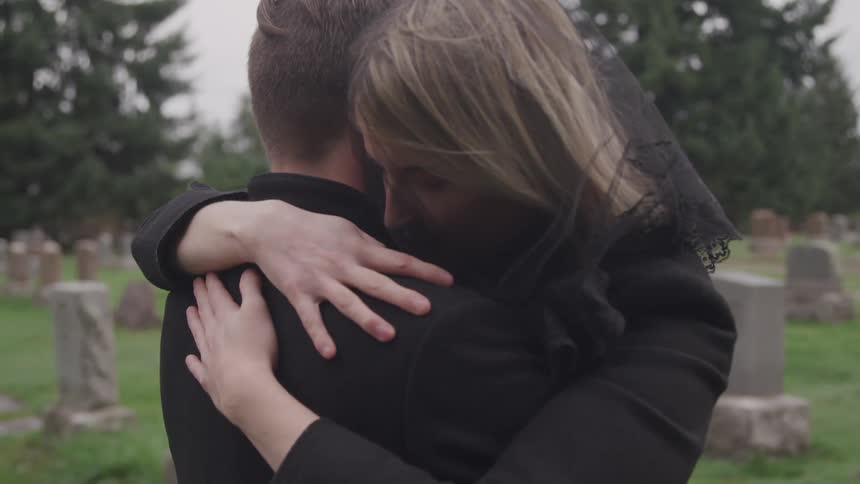 A man and woman dressed in all black embrace while standing in a cemetery during the day