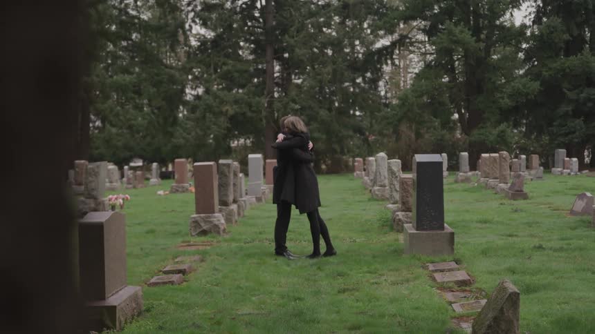 A man and woman embrace while standing between rows of graves in a graveyard during the day