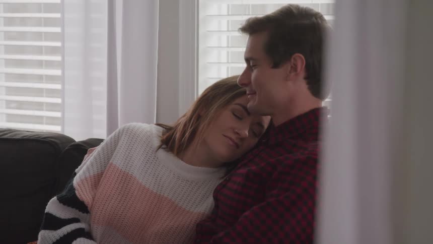 Man with a red striped shirt and woman with a sweater sitting on a sofa look into each others eyes with affection and hold each other