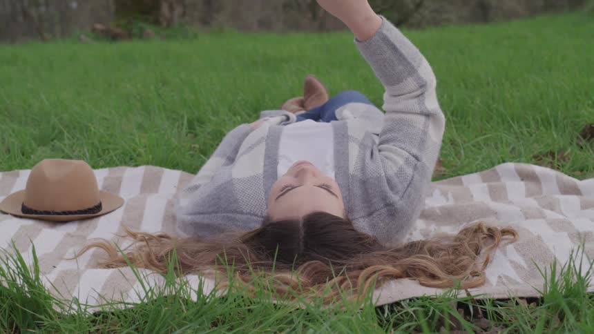 Woman with brown hair lies down on green grass and raises her right hand up in the air
