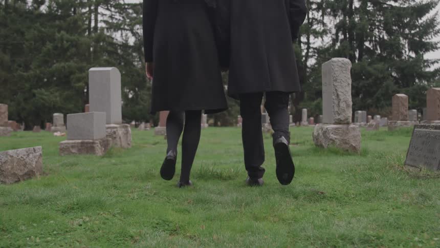 Male and female dressed in all black apparel walk side by side on the grass between the grave markers a cemetery