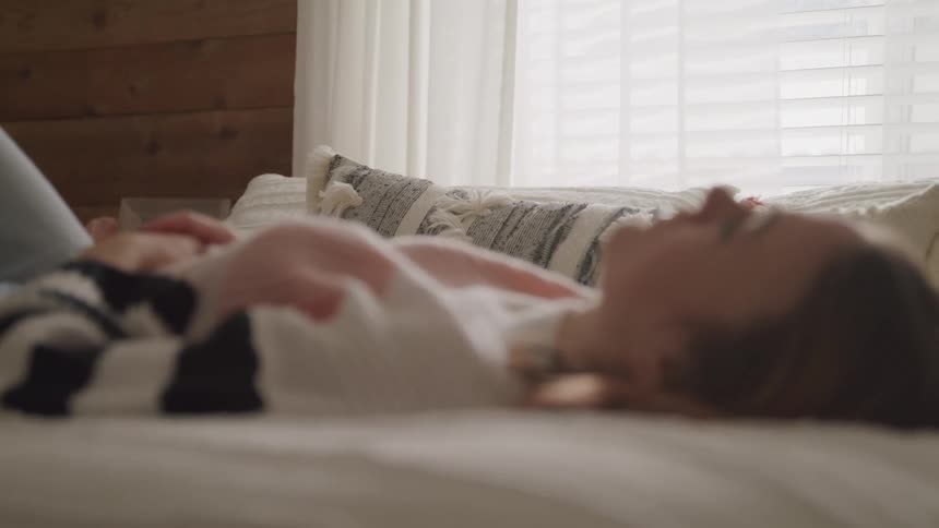 Woman with a sweater lies down on bed and looks up at the ceiling in bedroom