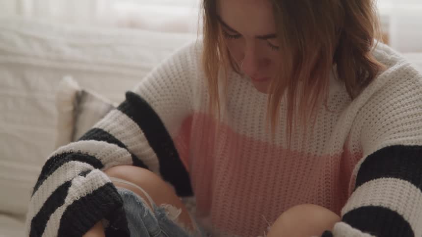 Woman with brown hair a loose knit sweater and ripped jeans sits on bed deep in thought and looks down